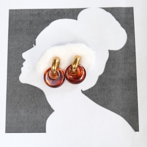 May include: A pair of hoop earrings featuring a polished gold-tone metal top section connected to a circular, translucent amber-colored resin base. The earrings have a smooth, glossy finish and a rounded, chunky design that creates a distinct contrast.