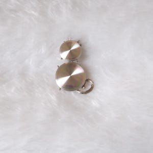 May include: A silver-colored cat-shaped brooch. The brooch is made of shiny metal and features a circular head and body, with a small curled tail. The brooch is set against a white, fluffy background.