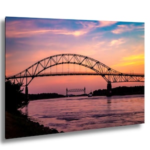 May include: A scenic photograph of a bridge silhouetted against a vibrant sunset. The sky transitions from pink to orange and blue, reflecting in the water below. The bridge's intricate metalwork is a prominent feature.