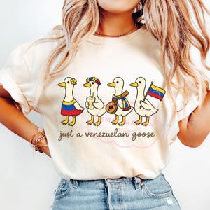 May include: Cream-colored t-shirt featuring a whimsical illustration of four cartoon geese, each adorned with Venezuelan-themed accessories. The geese are dressed in outfits with the colors of the Venezuelan flag. The text "just a venezuelan goose" is printed below the geese.