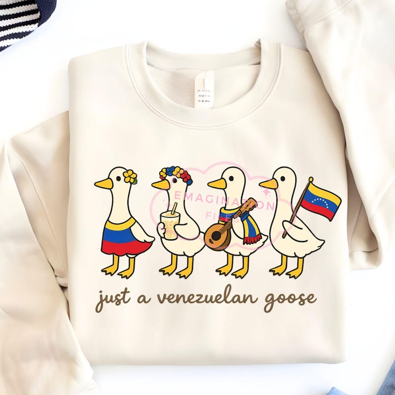 May include: Cream-colored sweatshirt featuring four cartoon geese, each adorned with Venezuelan-themed accessories. One goose wears a flower crown and a dress in the colors of the Venezuelan flag. The text "just a venezuelan goose" is printed below the geese.