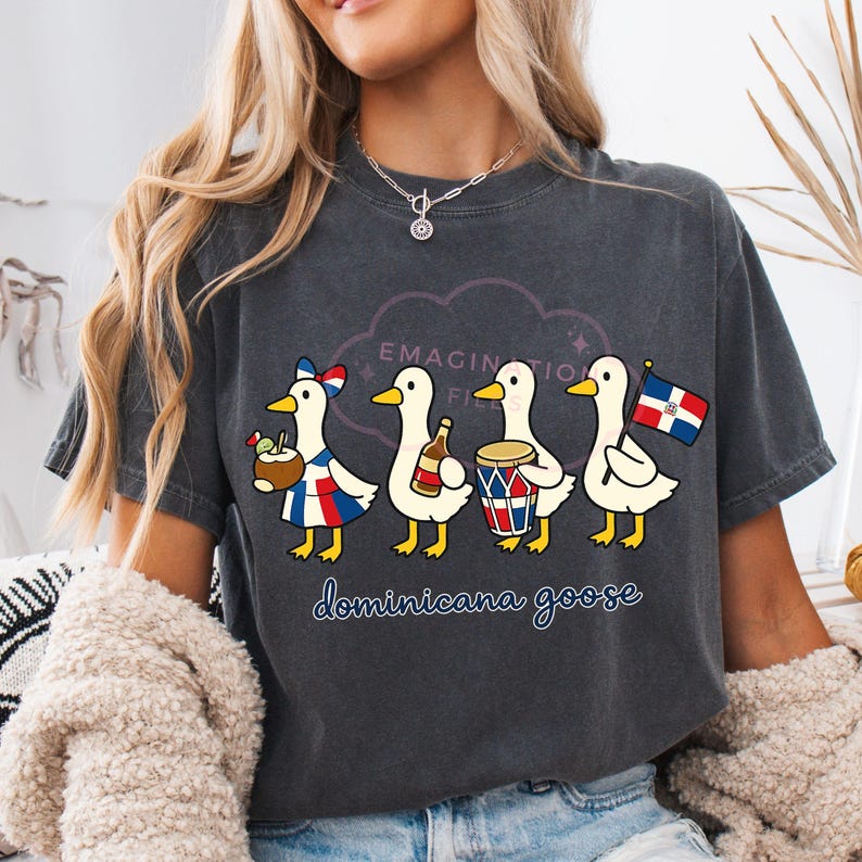 May include: Dark grey t-shirt with a cartoon design of four geese in Dominican Republic outfits. The geese are holding a drink, a bottle, a drum, and a flag. The shirt has the text "EMAGINATION FI" and "dominicana goose".