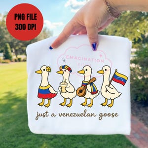 May include: White sweatshirt featuring four cartoon geese, each adorned with Venezuelan-themed accessories. The geese are wearing a flower crown, holding a drink, playing a mandolin, and holding a Venezuelan flag. Text reads "just a venezuelan goose."