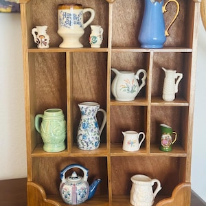 Puede incluir: Una estantería de madera que exhibe varias jarras, tazas y cremeras de cerámica. Los artículos presentan diferentes colores y diseños, incluyendo diseños florales y las palabras "Home Sweet Home" en una taza. La estantería es de madera marrón claro.