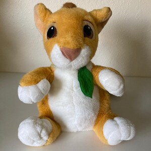 May include: A plush lion cub toy with orange and white fur, large brown eyes, and a green leaf accent. The stuffed animal is sitting upright with its paws forward.