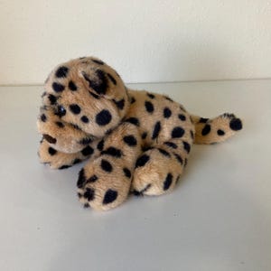May include: A plush toy cheetah in a lying position. The cheetah has a tan body with black spots and dark eyes. The toy is made of soft, fuzzy material.