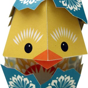 May include: A paper craft chick hatching from an egg. The egg is decorated with blue and white floral patterns. The chick is yellow with a black eye, orange beak, and white eyelashes.