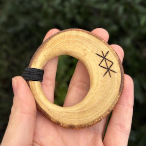 Op de afbeelding: Een houten ring met een gegraveerd zwart symbool. De ring is gemaakt van een natuurlijke houtschijf en heeft een gladde, gepolijste afwerking.