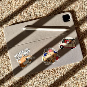 Puede incluir: Una funda de tableta gris claro con un lápiz óptico blanco y tres pegatinas con temática de perros. Una pegatina muestra un corgi, otra un perro durmiendo con tazas de café y la última una furgoneta con perros.