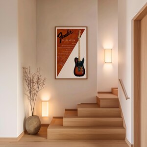 以下が含まれることがあります： Fender Telecasterギターが描かれた額入りのポスター。ポスターは茶色と白の背景にFenderのロゴとギターの詳細が描かれています。ギターは黒、茶色、オレンジ色です。ポスターは木製の階段の近くの壁に掛けられています。