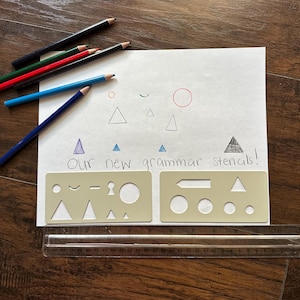 May include: A sheet of white paper with colorful drawings and the text "Our new grammar stencils!" is surrounded by colored pencils, two beige stencils, and a clear ruler. The stencils have various shapes.