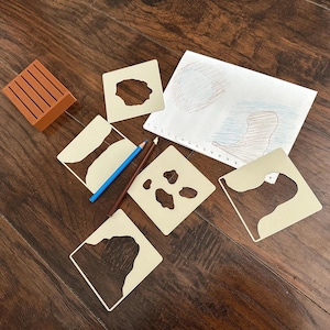 May include: An assortment of art supplies including stencils, colored pencils, and a small wooden block. The stencils are square with various cut-out shapes. A drawing with pencil and crayon is also present, suggesting a creative activity.