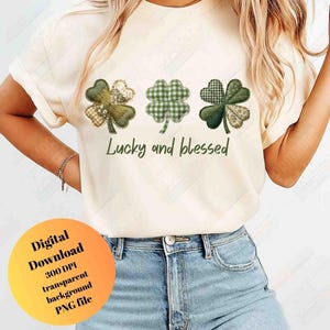 May include: Cream-colored t-shirt featuring three shamrock designs in shades of green and tan, with various patterns. The text "Lucky and blessed" is printed below the shamrocks. The shirt is paired with blue jeans.