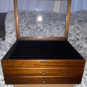 May include: A rectangular wooden jewelry box with a hinged glass lid and two lower drawers. The interior features black velvet lining with multiple slots for rings or small items. The exterior has a medium brown finish and small brass knobs.