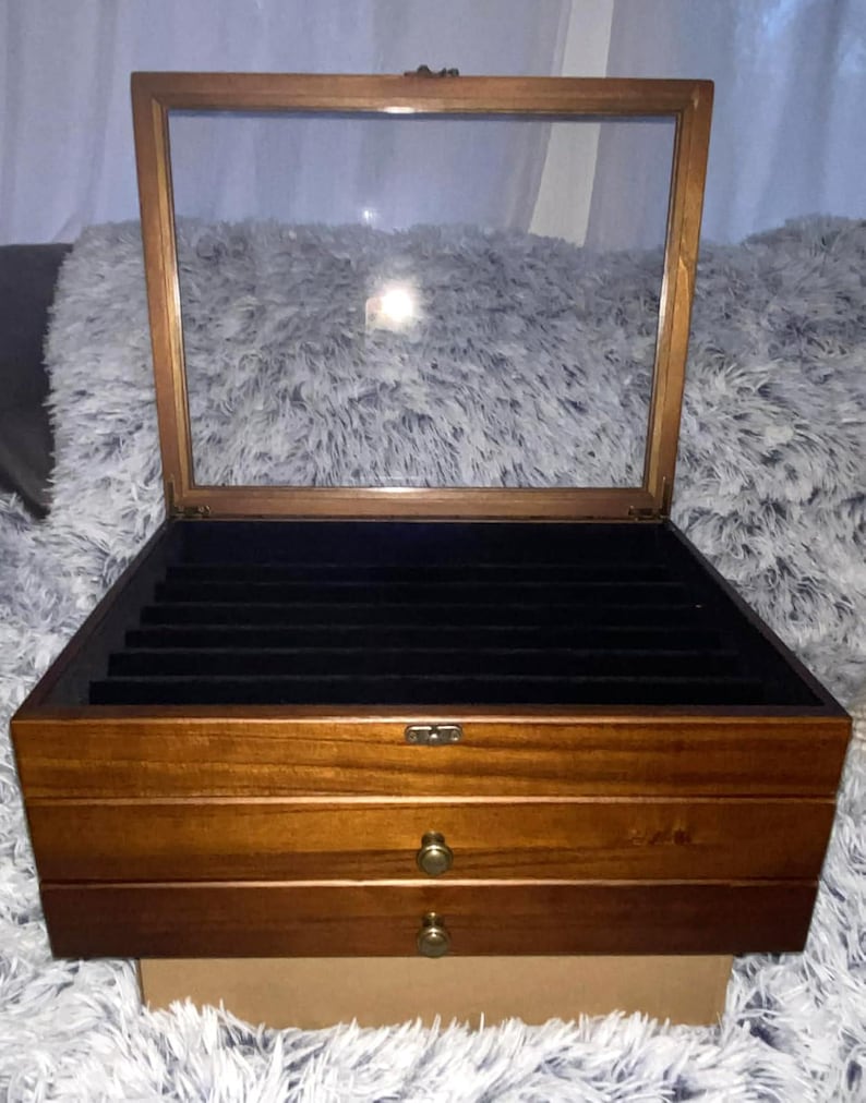 May include: A rectangular wooden jewelry box with a hinged glass lid and two lower drawers. The interior features black velvet lining with multiple slots for rings or small items. The exterior has a medium brown finish and small brass knobs.