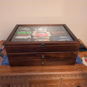 May include: A dark brown wooden display case with a glass top lid and two pull-out drawers. The glass lid reveals a collection of various folding pocket knives stored inside. The case features antique-style metal latches and small round drawer knobs.