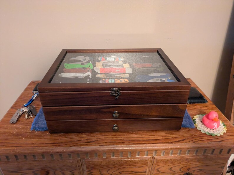 May include: A dark brown wooden display case with a glass top lid and two pull-out drawers. The glass lid reveals a collection of various folding pocket knives stored inside. The case features antique-style metal latches and small round drawer knobs.