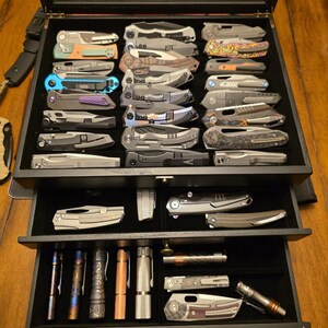 May include: A black wooden display case with three drawers containing a collection of folding pocket knives and cylindrical metal flashlights. The knives feature various metallic finishes, pocket clips, and handle textures, organized in rows.
