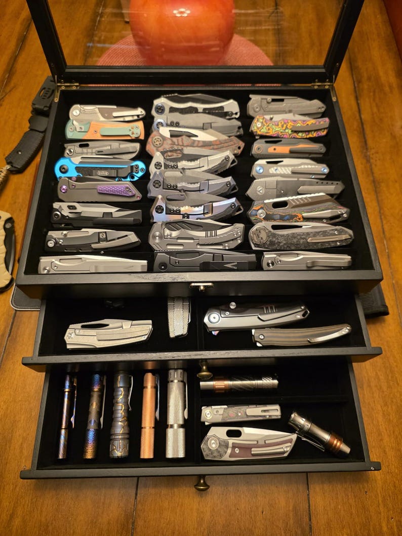 May include: A black wooden display case with three drawers containing a collection of folding pocket knives and cylindrical metal flashlights. The knives feature various metallic finishes, pocket clips, and handle textures, organized in rows.