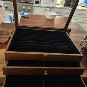 May include: A dark wood jewelry storage box with a hinged glass lid and two pull-out drawers. The interior is lined with black velvet, featuring multiple long slots in the top compartment and divided sections in the drawers with brass knobs.