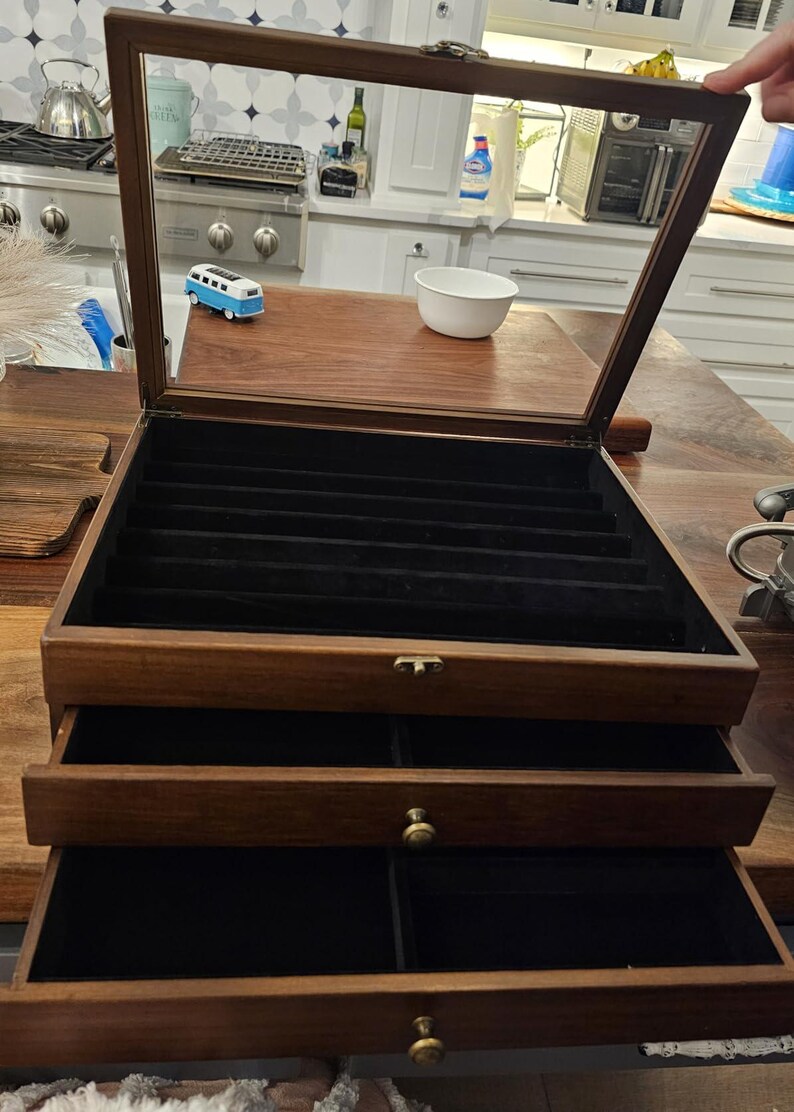 May include: A dark wood jewelry storage box with a hinged glass lid and two pull-out drawers. The interior is lined with black velvet, featuring multiple long slots in the top compartment and divided sections in the drawers with brass knobs.