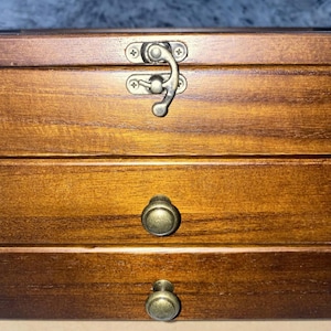 May include: A rectangular wooden storage box with a medium brown finish. The top section features a metal latch closure, while the two lower sections are drawers, each equipped with a round, antique-style brass knob for easy opening and access.