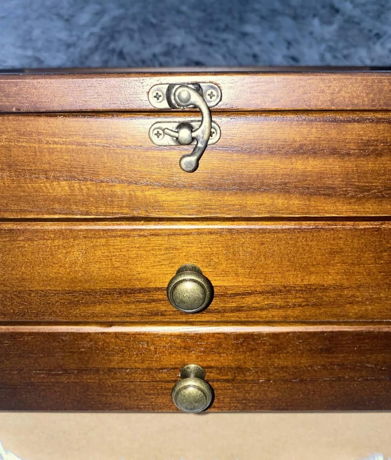 May include: A rectangular wooden storage box with a medium brown finish. The top section features a metal latch closure, while the two lower sections are drawers, each equipped with a round, antique-style brass knob for easy opening and access.
