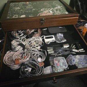 May include: A dark wood storage box with a glass top lid and a pull-out drawer. The interior is lined with black fabric and contains multiple coiled audio cables, various small electronic accessories, and clear plastic containers with ear tips.