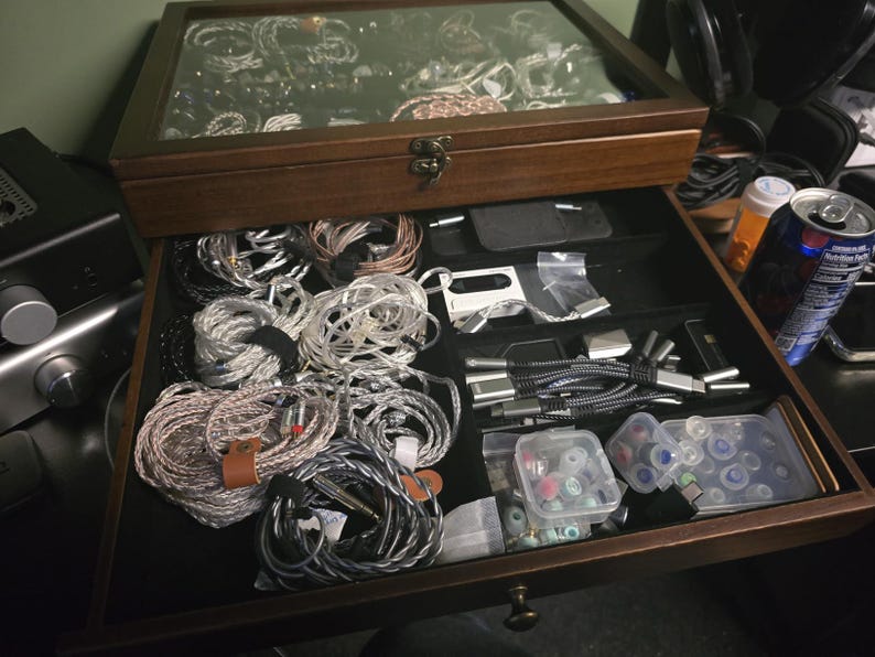 May include: A dark wood storage box with a glass top lid and a pull-out drawer. The interior is lined with black fabric and contains multiple coiled audio cables, various small electronic accessories, and clear plastic containers with ear tips.