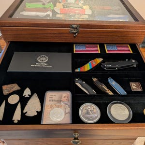 May include: A wooden display case with a glass top and an open drawer featuring black velvet lining. The drawer contains various items including pocket knives, two American flag patches, stone arrowheads, coins in protective cases, and a box labeled Quad Edge EDC Multitool Sharpener.