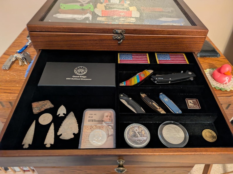 May include: A wooden display case with a glass top and an open drawer featuring black velvet lining. The drawer contains various items including pocket knives, two American flag patches, stone arrowheads, coins in protective cases, and a box labeled Quad Edge EDC Multitool Sharpener.