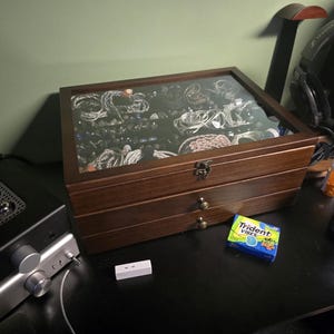May include: A rectangular wooden storage box with a glass lid, featuring two lower drawers with small round knobs and a metal latch. The interior is filled with various coiled cables and audio accessories, visible through the clear top panel.