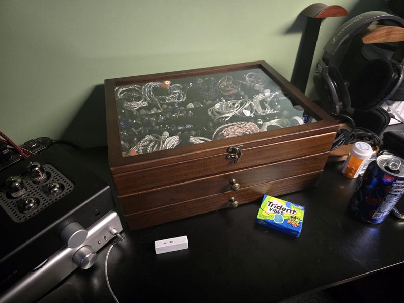 May include: A rectangular wooden storage box with a glass lid, featuring two lower drawers with small round knobs and a metal latch. The interior is filled with various coiled cables and audio accessories, visible through the clear top panel.
