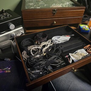 May include: A dark wood storage drawer with a black velvet interior, filled with various coiled audio cables, adapters, and small electronic accessories. The drawer features a small brass knob and is part of a larger wooden organizer unit.