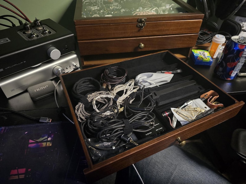 May include: A dark wood storage drawer with a black velvet interior, filled with various coiled audio cables, adapters, and small electronic accessories. The drawer features a small brass knob and is part of a larger wooden organizer unit.