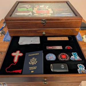 May include: A dark wood display case with a glass top lid and a pull-out drawer. The drawer contains a United States passport, a crocheted cross, a car key fob, several circular patches, a small cleaning cloth, and a rectangular wooden tag.