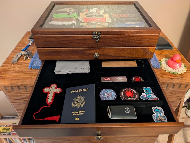 May include: A dark wood display case with a glass top lid and a pull-out drawer. The drawer contains a United States passport, a crocheted cross, a car key fob, several circular patches, a small cleaning cloth, and a rectangular wooden tag.