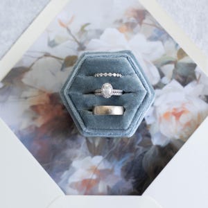 May include: Three silver rings in a blue velvet ring box. The rings are stacked on top of each other. The top ring has a large oval-shaped diamond.