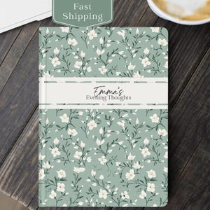 May include: A journal with a floral pattern in white and green. The journal has the text "Emma's Evening Thoughts" on a white band across the front. The background is a muted green color. The journal is on a dark wooden surface.