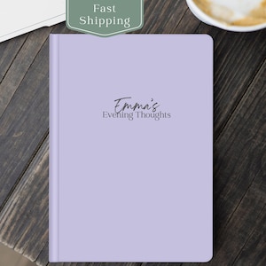 May include: A lavender-colored journal with the text "Emma's Evening Thoughts" in a script font. The journal is closed and has a smooth cover. The background is a dark wood surface.