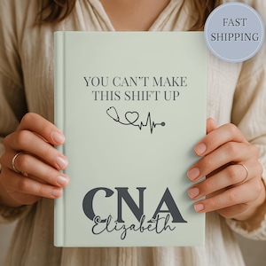 May include: A light green hardcover journal featuring the dark gray text "YOU CAN'T MAKE THIS SHIFT UP" above a black stethoscope and heart EKG line graphic. Below this, the letters "CNA" are printed in large dark gray font with the name "Elizabeth" below.