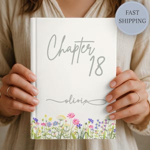 May include: A white journal with the text "Chapter 18" and the name "olivia" in gray script. The bottom edge features a watercolor floral design with pink, blue, and yellow flowers. A round sticker with "FAST SHIPPING" is in the upper right corner.