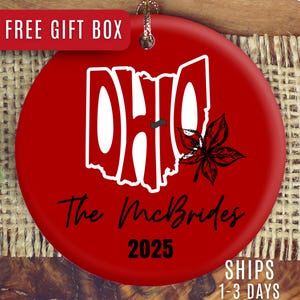 May include: A red ceramic ornament with a white outline of Ohio and the word "OHIO" in white. The ornament also features a black buckeye leaf and the text "The McBrides 2025". A gold string is attached for hanging.