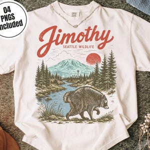 May include: A white t-shirt featuring a graphic print with the text "Jimothy SEATTLE WILDLIFE". The design depicts a raccoon walking near a river, a forest of pine trees, a mountain, and a red sun. The text "04 PNGS INCLUDED" is in a circle.
