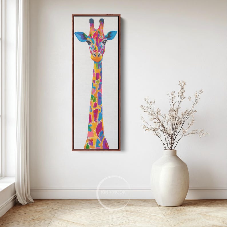 May include: A vibrant painting of a giraffe, framed in dark wood, against a white backdrop. The giraffe features a colourful pattern of blues, pinks, yellows, and oranges. The artwork is displayed on a white wall, with a vase of dried flowers in the foreground.