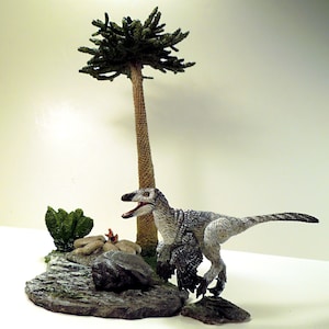 Może przedstawiać: Figurkę dinozaura w kolorze szarym i białym z długim ogonem i otwartą paszczą, stojącą na skalistym podłożu z małym zielonym krzewem i brązową palmą.