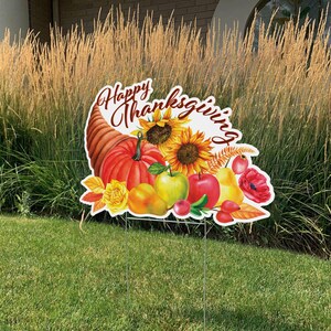 May include: Thanksgiving yard sign featuring a cornucopia, pumpkins, sunflowers, apples, and pears. The sign has the words "Happy Thanksgiving" in a decorative script. The sign is supported by a metal stake and is placed on a green lawn.