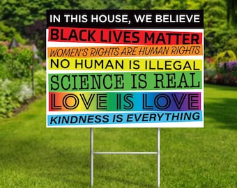 May include: A yard sign with a rainbow background and black text. The text reads: "In this house, we believe Black Lives Matter, Women's Rights are Human Rights, No Human is Illegal, Science is Real, Love is Love, Kindness is Everything."