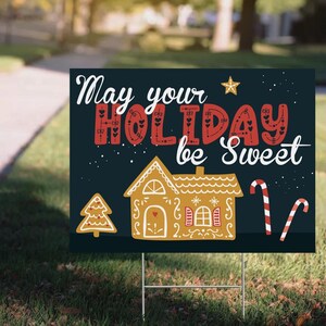 May include: A holiday yard sign with the message "May your HOLIDAY be Sweet" in white and red lettering. The sign features a gingerbread house, candy canes, and a Christmas tree on a dark blue background.