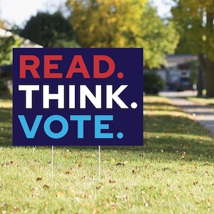May include: A rectangular yard sign with a dark blue background featuring the text READ in red, THINK in white, and VOTE in light blue, each followed by a period. The sign is supported by two thin metal stakes inserted into the ground.